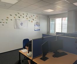 Schulungsraum mit mehreren Arbeitsbereichen, die durch blaue Trennwände voneinander abgegrenzt sind und mit 2 PC-Bildschirmen, Tastatur, Maus und Headset ausgestattet sind. Fensterreihe und gebastelte Schmetterlinge an der Wand.