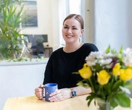 Frau mit braunem Haar sitzt am Tisch und hält eine blaue Tasse. Vor ihr steht ein Strauß Blumen. Im Hintergrund befinden sich Arbeitsplätze und Pflanzen.