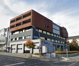 Außenansicht des Gebäudes. Im Erdgeschoss befinden sich mehrere Garagen und auf der grauen Fassade ist ein rotes Kreuz zu sehen. Die darüberliegenden Etagen haben eine schwarze und rote Fassade sowie viele Fenster und Balkone.