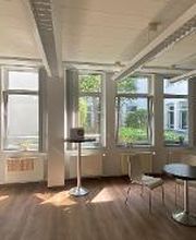 Pausenraum mit mehreren Stühlen und Tischen vor einer roten Wand. Links eine weiße Küche mit Spüle, Kühlschrank und Mikrowelle.