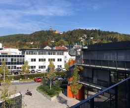 Sonnige Aussicht vom Balkon mit Blick auf einen Hof mit Grünflächen und Bäumen. Im Hintergrund ein weißes Nachbarhaus und Parkplätze hinter dem ein Berg mit Wald ragt.