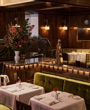White-clothed restaurant tables with green velvet banquettes positioned near a dark marble ledge featuring a large, colorful floral arrangement in a tall vase.