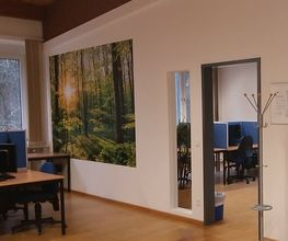 Ein PC-Arbeitsplatz, ein Bild einer Waldszene und eine Tür in einen anderen Raum, in dem sich ebenfalls PC-Arbeitsplätze befinden.