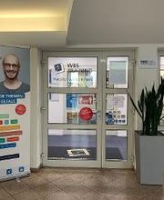 Innenansicht des Eingangsbereiches. Eine Glastüre mit der Aufschrift WBS TRAINING Herzlich willkommen. Davor steht ein Werbeaufsteller, ein Aufsteller mit Infobroschüren sowie eine große Pflanze.