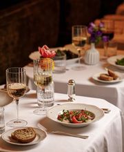 Elegant restaurant table with a refined seafood dish—thinly sliced fish or shellfish dressed with herbs and tomatoes—served on a white-rimmed plate, accompanied by crusty bread, wine glasses, soft candlelight, and delicate floral arrangements on crisp white linens.