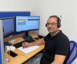 Ein Teilnehmer sitzt an seinem Arbeitsplatz vor 2 PC-Bildschirm und trägt sein Headset. Er macht sich Notizen und schaut gut gelaunt in die Kamera.