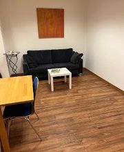 Eine schwarze Couch in einem Pausenraum. Am Bildrand ist ein Holztisch mit einem weiteren Stuhl zu sehen.