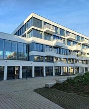 Weißes Gebäude mit mehreren Fenstern und Balkonen. Davor Grünflächen und Pflastersteine