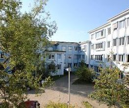 Ein großes Gebäude mit weißer Fassade und hellblauen Fensterrahmen. Es ist ein sonniger Tag, der Himmel ist blau und es sind grüne Baumkronen im Bild.