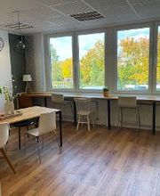 Ein großer Holztisch mit Stühlen im Aufenthaltsraum. Ein weißes Bücherregal und Fensterfront mit Blick ins Grüne sowie weiteren Sitzgelegenheiten.