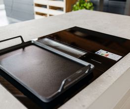 Das Bild zeigt ein modernes Induktionskochfeld mit integriertem Muldenlüfter auf einer hellen Kücheninsel. Auf der linken Kochzone liegt eine hochwertige Grill- bzw. Teppanyaki-Platte mit seitlichen Griffen, die vielseitige Zubereitungsmöglichkeiten direkt auf dem Kochfeld ermöglicht. Die helle Arbeitsplatte bildet einen eleganten Kontrast zur schwarzen Glaskeramikfläche. Der Hintergrund ist dezent unscharf und lenkt den Fokus auf die innovative Kochtechnik.