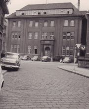 Eine historische Aufnahme des Rathauses II in Oldenburg mit Blick auf die gepflasterte Straße und parkende Autos aus vergangenen Jahrzehnten. Das Bild vermittelt den Charme des alten Stadtbildes.