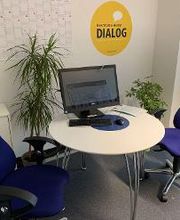 Ein kleiner Beratungsraum mit einem runden weißen Tisch, darauf ein Monitor, Tastatur und Maus. Zwei blaue Bürostühle stehen am Tisch. An der Wand ein gelber Kreis mit „Beratung heißt Dialog“, daneben ein Kalender und Pflanzen im Raum.