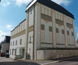 Außenansicht vom Gebäude und Eingang. Das Gebäude hat eine weiße Fassade mit einem gelben Muster.
