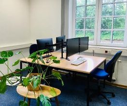 Zwei Arbeitsplätze im Schulungsraum mit zwei Schreibtischen, je 2 Bildschirmen und blauen Bürostühlen vor einem großen zweiflügeligen Sprossenfenster. Davor Tisch mit Grünpflanze. Weiße Wände, blauer Teppich.