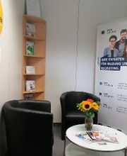 Gemütliche Sitzecke mit 2 schwarzen Ledersesseln, einem weißen Beistelltisch auf dem ein Blumenstrauß steht sowie ein Werbeaufsteller und Holzregal im Hintergrund.