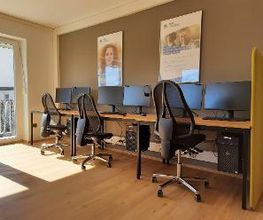 Schulungsraum mit 3 Arbeitsplätzen mit jeweils 2 Bildschirmen, einem Bürostuhl, Tastatur, Maus und Headset. Darüber hängen 2 WBS TRAINING Plakate, daneben eine gelbe Trennwand.