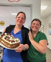 Bettina Henri (links) und Denise Kir (rechts), Mitarbeiterinnen der WBS TRAINING, halten lachend eine Torte im Büro. Bettina hält die Torte, während Denise sie umarmt. Beide strahlen Freude aus und vermitteln die herzliche Atmosphäre des WBS Teams.