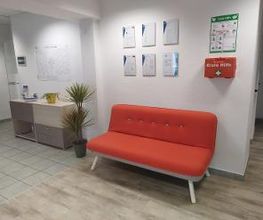 rotes Sofa, daneben ein Regal und Pflanzen. Über dem Sofa mehrere Plakate und ein Erste Hilfe Koffer und Instruktionen.