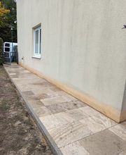 Schmaler Gartenweg entlang eines Wohnhauses, ausgeführt mit hellen Natursteinplatten. Krippner, Garten- und Landschaftsbau bei Berlin schafft mit solchen Pflasterarbeiten sichere und optisch ansprechende Verbindungen zwischen Garten, Eingang und Terrasse.