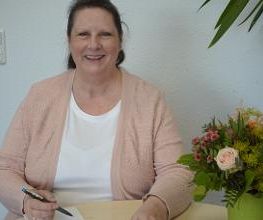 Eine Frau in rosa Strickjacke sitzt an einem Tisch und hält einen Stift über ein Notizbuch. Sie lächelt in die Kamera. Rechts auf dem Tisch steht ein Blumenstrauß in einer grünen Vase. Im Hintergrund sind eine weiße Wand und eine Pflanze zu sehen.
