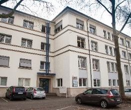 Außenansicht auf das Gebäude von der gegenüberliegenden Straßenseite. Hier wird der Eingang präsentiert. Es gibt einige Parkplätze vor dem Gebäude.