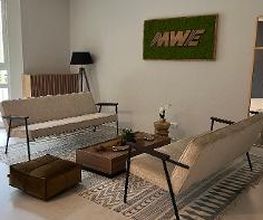 Foyer mit 2 modernen und stilvollen Sofas sowie einem braunen Beistelltisch und einem braunen Sitzkissen. Im Hintergrund eine Stehlampe sowie eine grüne Zimmerpflanze vor einem großen Fenster.