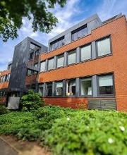 Außenansicht des Schulungsgebäude mit orangenen Klinkern und grauer Fensterumrahmung. Das Gebäude ist umgeben von grünen Pflanzen und Bäumen.