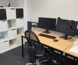 Arbeitsplatz bestehend aus 2 PC-Bildschirmen, Tastatur, Maus und Headset sowie einem schwarzen Bürostuhl. Daneben ein Regal mit mehreren Ablagefächern. 