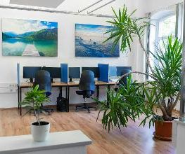 Schulungsraum mit 3 Arbeitsplätzen mit jeweils 2 Bildschirmen, einem Bürostuhl und Tastatur und Maus. Darüber hängen 2 Landschaftsbilder. Hinter den Tischen stehen Pflanzen.