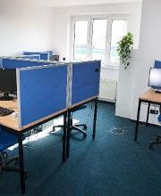 Dieses Bild zeigt einen Schulungsraum. Zu sehen sind Schreibtische, welche jeweils mit zwei Bildschirmen, einer Tastatur, Maus und Headset ausgestattet sind.