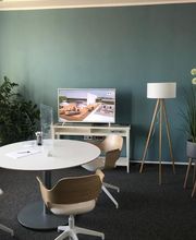 Aufenthaltsraum mit blauer Wand. Davor ein Regal mit TV. Daneben eine Stehlampe und mehrere Pflanzen. Im Raum ein Tisch mit 3 Stühlen