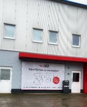 Außenansicht der Schulungsstätte mit Beschilderung der dort sitzenden Unternehmen. Graue Fassade und rote Überdachung,
