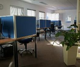 Schulungsraum mit mehreren Arbeitsbereichen, die durch blaue Trennwände voneinander abgegrenzt sind und mit PC-Bildschirmen, Tastatur, Maus und Headset ausgestattet sind. Fensterreihe und Bilder an der Wand.
