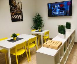Modern und hell gestalteter Aufenthaltsbereich mit weißen Tischen und gelben Stühlen. Ikea-Kallax Raumteiler. Blumendeko, große Pflanze in der Ecke, Gesellschaftsspiele, Bücher im Regal. Wandbild.