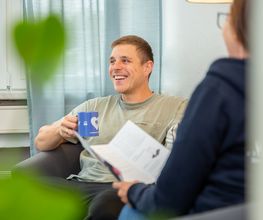 Mann sitzt in einem Sessel und hält eine blaue Tasse in der Hand. Ihm gegenüber sitzt eine Frau mit einem Flyer in der Hand.