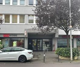 Eingangsbereich eines Bürogebäudes, dvor eine Straße und ein geparktes Auto. Links im Erdgeschoss befindet sich eine Apotheke.
