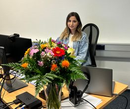 Eine junge Frau sitzt an einem PC. Vor ihr steht ein großer Blumenstraß in einer Vase.