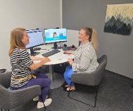 Eine Beratungssituation. Zwei Frauen sitzen an einem Tisch vor 2 Bildschirmen und unterhalten sich. Hellgraue Wand mit Textildekoration.
