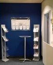 Eingangsbereich mit einer gelben und einer blauen Wand, vor der ein Bistrotisch und zwei Werbeaufsteller stehen.