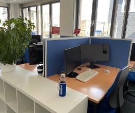 Arbeitsplätze mit blauen Trennwänden, 2 Bildschirmen, Tastatur, Maus und Headset. Der Raum hat viele Fenster. Es befindet sich ein weißes Regal auf dem eine grüne Topfpflanze und eine Getränkeflasche steht in dem Raum.