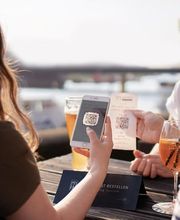 Zwei Frauen sitzen in einem Außenbereich und scannen mit ihren Smartphones einen QR-Code, während sie Getränke genießen. Das Bild zeigt die nahtlose Integration digitaler Technologien von gastronovi wie QR-Code-Bestellsysteme zur Selbstbedienung in der Gastronomie.