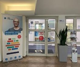Innenansicht des Eingangsbereiches. Eine Glastüre mit der Aufschrift WBS TRAINING Herzlich willkommen. Davor steht ein Werbeaufsteller, ein Aufsteller mit Infobroschüren sowie eine große Pflanze.