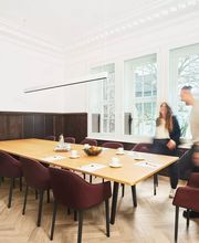 Ein stilvoller Meetingraum in der Alten Stadtkasse Oldenburg, ausgestattet mit einem großen Konferenztisch und eleganten Polsterstühlen. Helle Fensterfronten sorgen für eine angenehme Atmosphäre, während dunkle Holzelemente den historischen Charme des Raumes unterstreichen. Perfekt für Tagungen, Seminare und Geschäftstreffen mit bis zu 12 Personen.