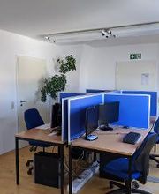 Raum mit 4 Arbeitsplätzen und 2 Türen. Arbeitsplätze mit jeweils 2 Bildschirmen, einem Bürostuhl, Tastatur und Maus. Links und rechts hängen Plakate der WBS GRUPPE und WBS TRAINING. 