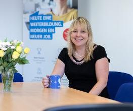 Eine Frau mit blonden Haaren und schwarzer Bluse sitzt an einem Tisch. Sie hält eine blaue Tasse in der Hand.