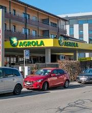 AGROLA Tankstelle in Zug; auf linker Seite Tankstelle hinter geparkten Autos; grosses Totem mit AGROLA Logo und digitalen Preisschildern sowie Selfwash Logo