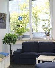 Wartebereich mit Blick auf grüne Bäume. Ein blaues Sofa steht vor dem Fenster mit Pflanzen auf dem Fensterbrett. Rechts steht ein Flyerständer, links hängt ein TV, darunter eine Sitzbank mit bunten Kissen. 