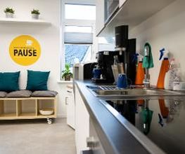 Voll ausgestattete Küchenzeile. Kleine Sitzbank mit Kissen, darüber hängt ein gelber, runder Aufkleber mit der Aufschrift Mach einfach mal Pause und einer weißen Kaffeetasse.