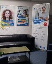 Aufenthaltsbereich mit Ledercouch und Sesseln, einer großen, grünen Zimmerpflanze sowie Druckstation. Zusätzlich steht ein Werbeaufsteller im Bereich. 